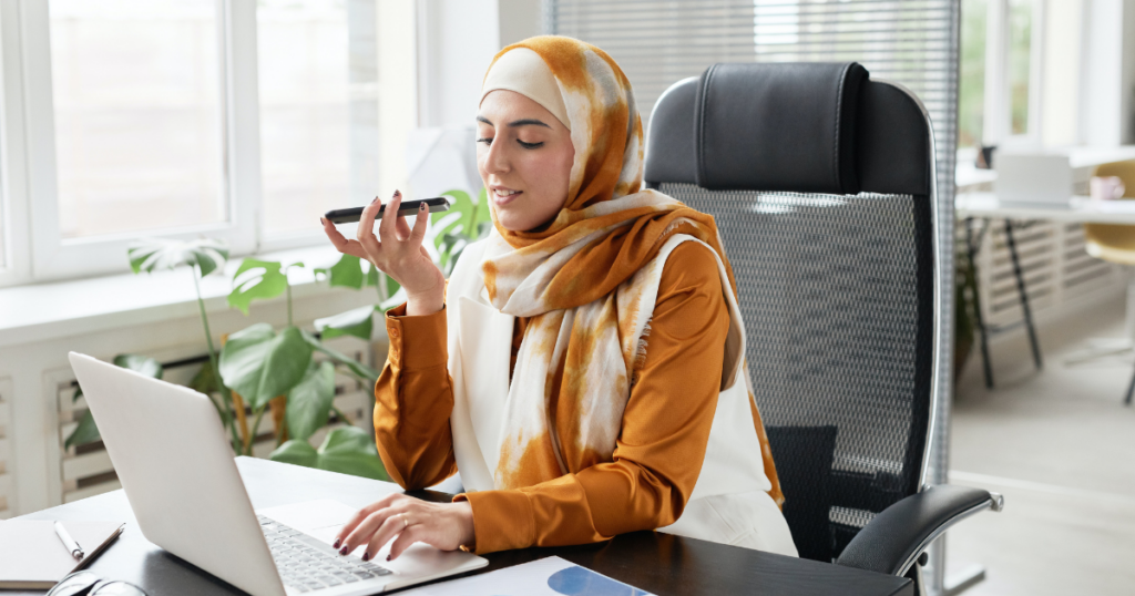 A woman wearing a hijab is speaking into the phone at her desk
